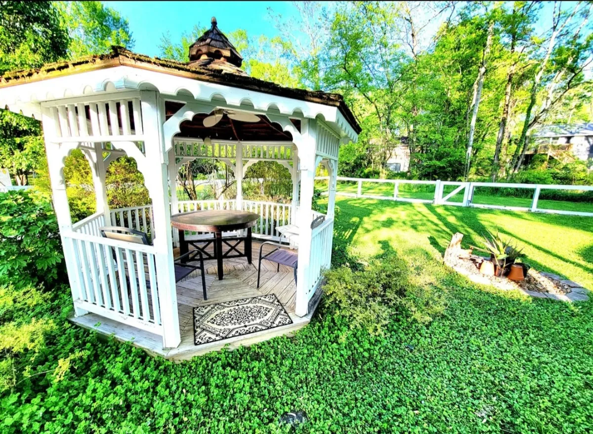 A peaceful exterior scene featuring a gazebo surrounded by lush greenery. There is a decorative rug, table, and chairs set out, inviting guests to relax with a coffee or enjoy quiet conversation while immersed in nature.