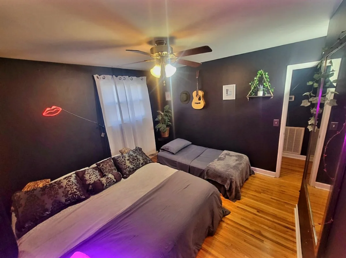 A large, cozy bed with decorative pillows is the center of this room, which features a soft neon lip light and a guitar mounted on the wall. The subdued lighting and casual decor create a relaxing, artistic environment ideal for unwinding after a busy day.