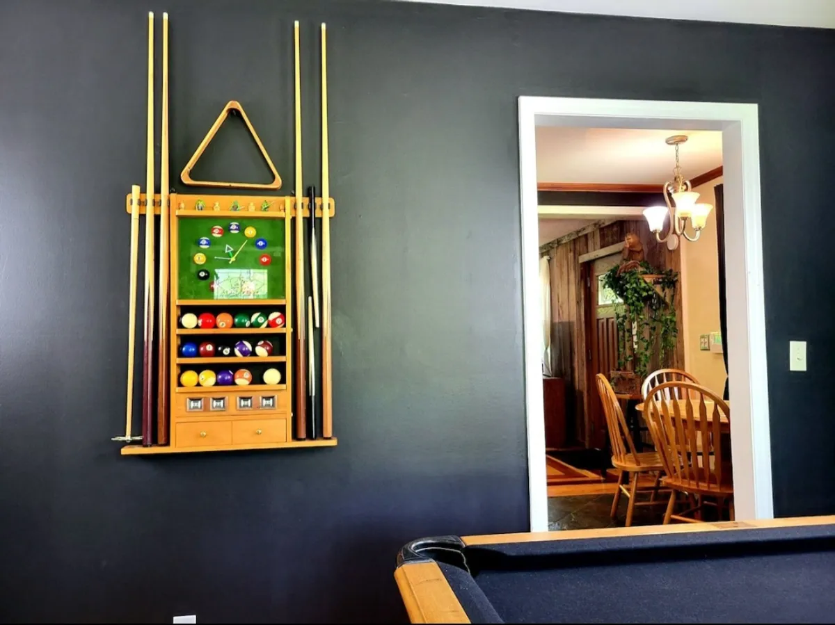A bright interior shot featuring a stylish pool table as the central focus. The wall-mounted cue rack and visible dining area through the doorway create a sense of flow and openness. The casual atmosphere is enhanced by the possibility of friendly competition and easy transition to shared meals, with space for spectators and participants alike.