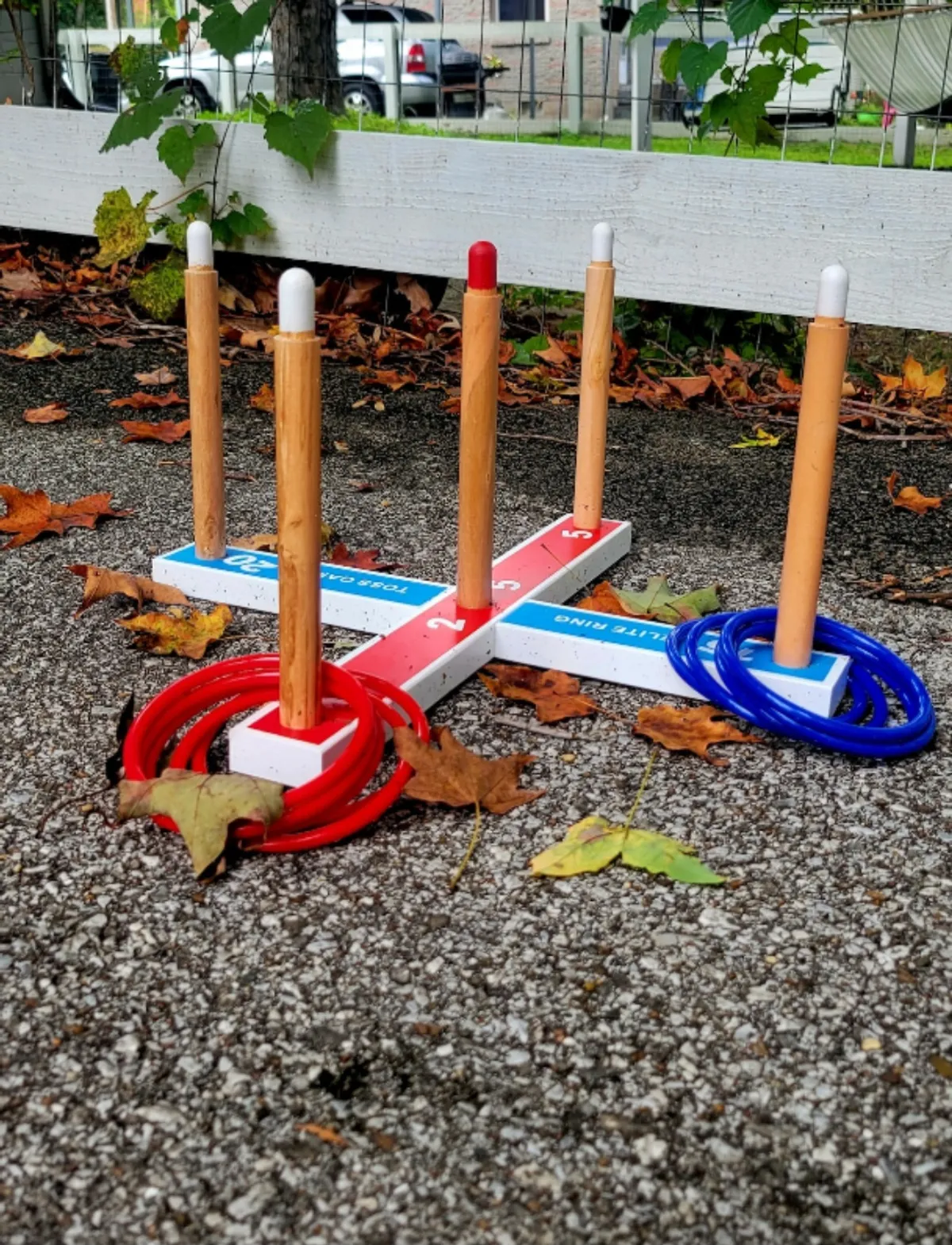 This image shows a nostalgic ring toss game set up outdoors, with autumn leaves scattered on the ground and a wooden fence in the background. The scene evokes a sense of safe, playful family fun in a backyard setting.