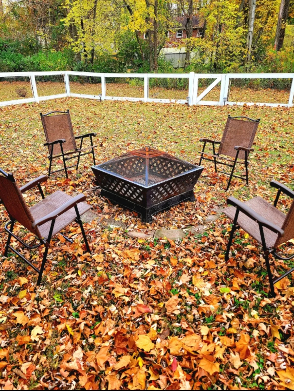A cozy outdoor fire pit area surrounded by chairs and autumn leaves within a fenced yard. The setting is intimate, perfect for evening conversations, sipping hot beverages, and watching sunsets together.