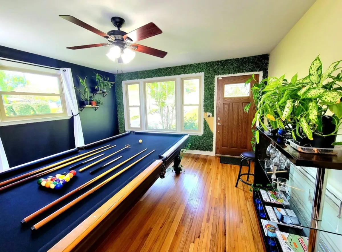 A relaxed, inviting game area with a pool table centered in the room, accented by greenery and large windows that flood the space with natural light. The arrangement encourages both competition and casual interaction, with enough room for onlookers to gather and cheer.
