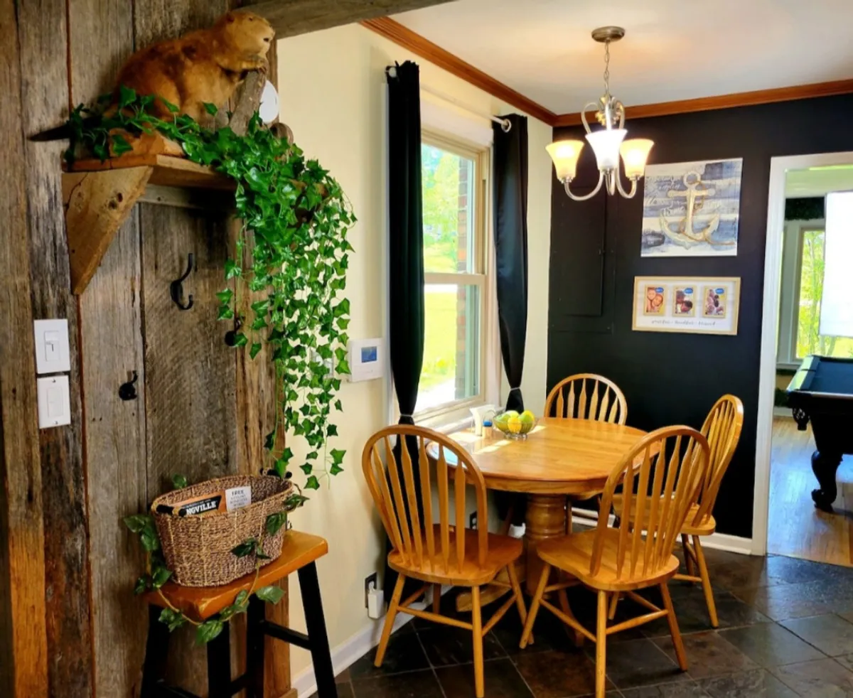 A warm and welcoming wooden dining table surrounded by greenery, with artwork on the wall and a stylish hanging light fixture above. The space evokes a feeling of coming together for meals, games, or celebrations.
