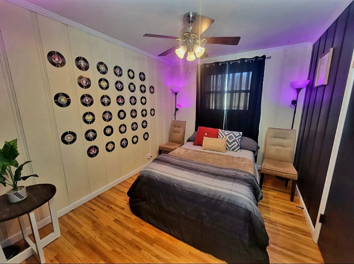 This bedroom features a bed adorned with multiple pillows and a wall decorated with vinyl records, flanked by two inviting chairs. The creative, nostalgic vibe adds character while offering a tranquil space for guests to relax, read, or listen to music.