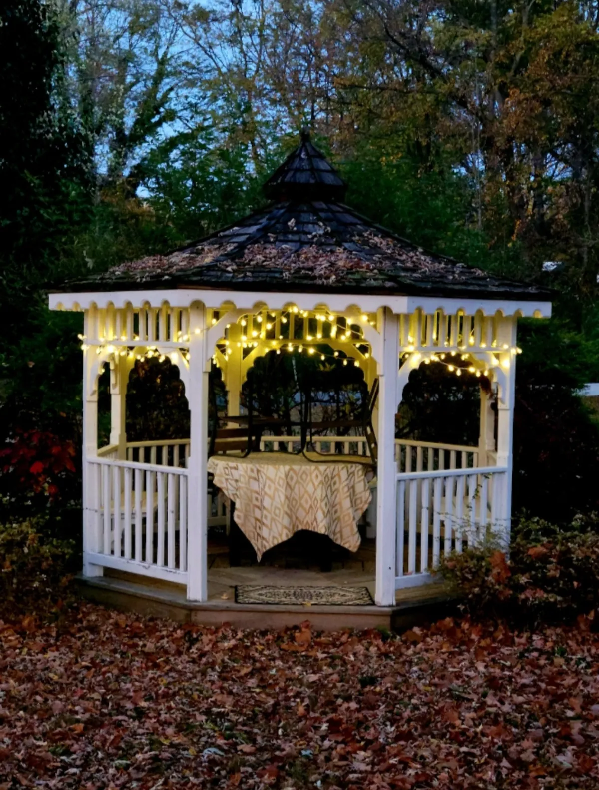 An exterior view of a cozy gazebo illuminated by twinkling string lights, with a table invitingly set for conversation. The surrounding autumnal landscape adds to the warm, communal atmosphere—ideal for evening gatherings with friends.