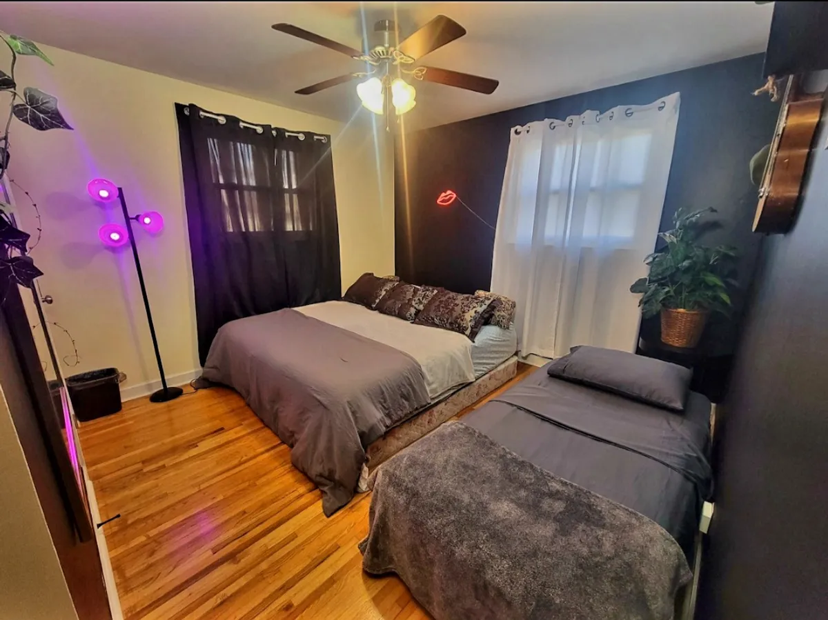 This image shows a bedroom with two comfortable beds, accented by a playful neon lips light and a decorative plant. The room is bright and inviting, with a colorful floor lamp adding warmth and personality. The arrangement suggests a space perfect for families or children to relax and enjoy restful moments together.