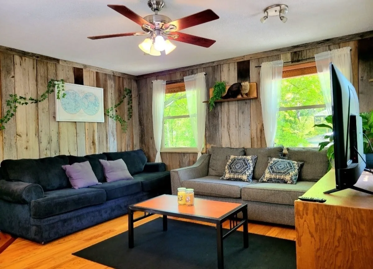 The image showcases a warm, inviting living room with plush couches arranged around a coffee table, a large TV perfect for movie nights, and a bright window offering a glimpse outside. The space is designed for comfort and togetherness, ideal for relaxing, entertaining, or unwinding with a book.